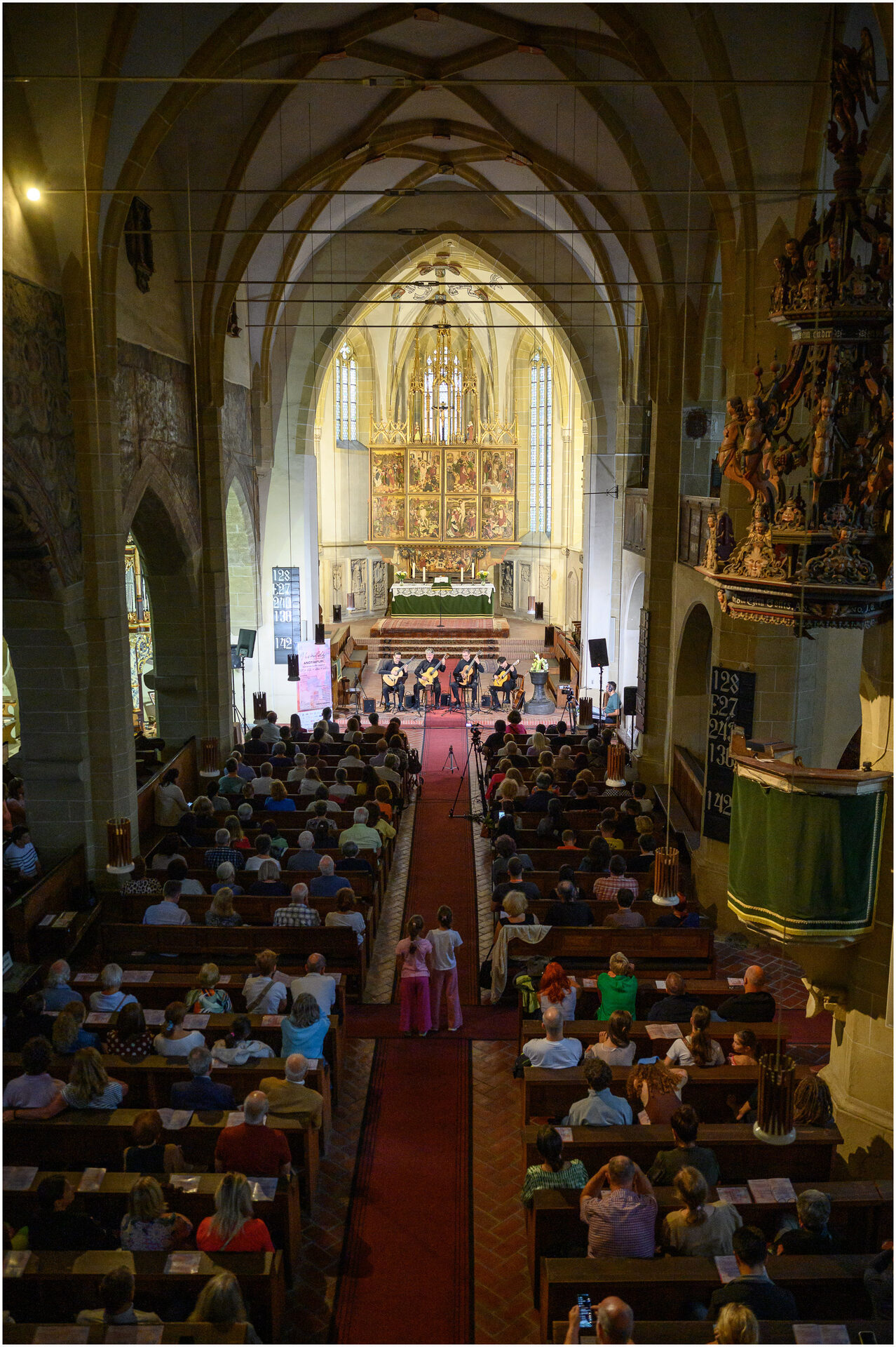 Vedere de ansamblu a interiorului unei biserici gotice, cu publicul urmărind concertul cvartetului de chitare lângă altar.