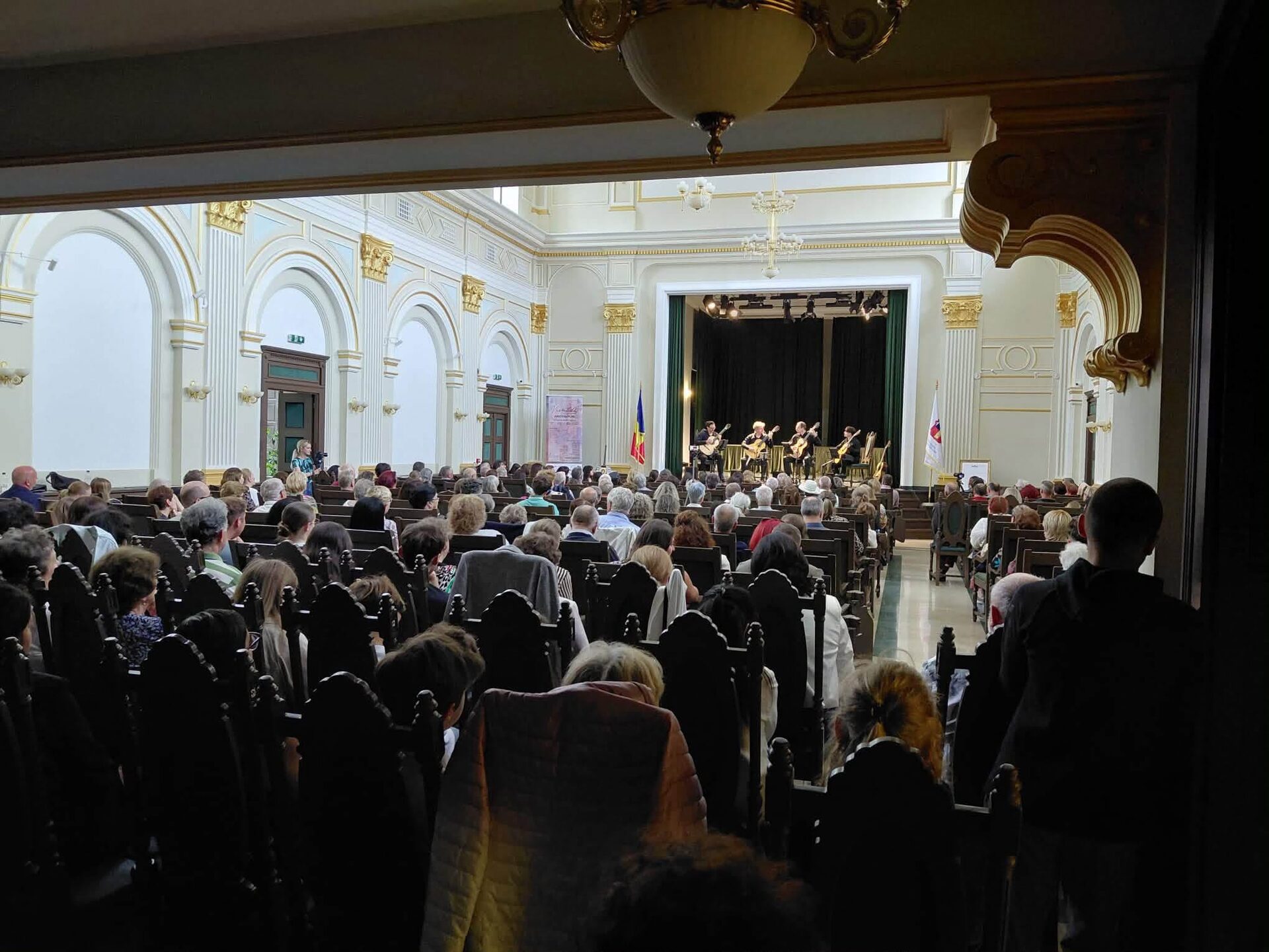 Publicul urmărind concertul într-o sală somptuoasă cu coloane și tavan auriu.