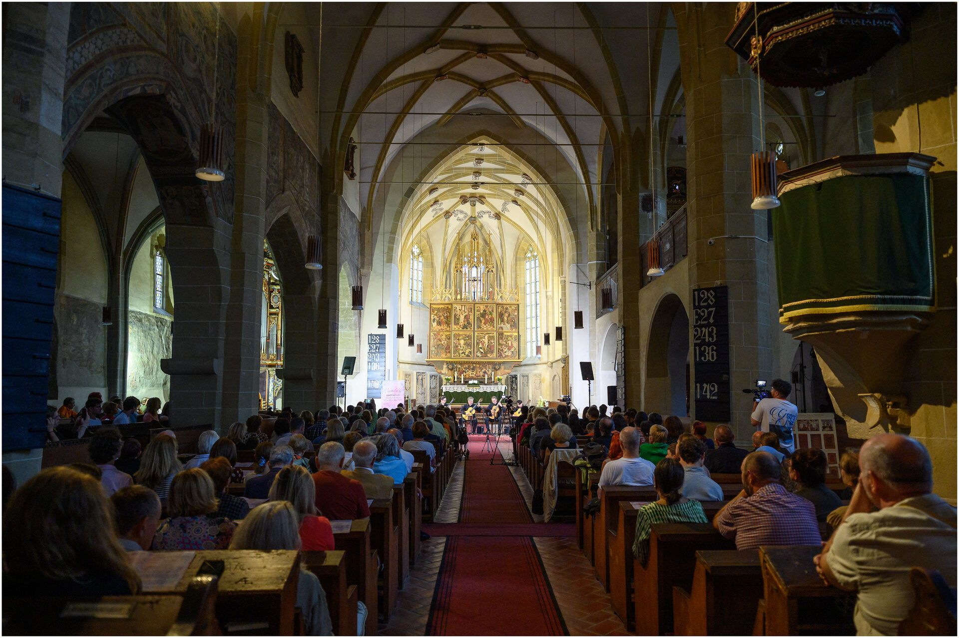 Vedere de sus a interiorului unei biserici gotice, cu publicul așezat pe strane și cvartetul cântând lângă altar.